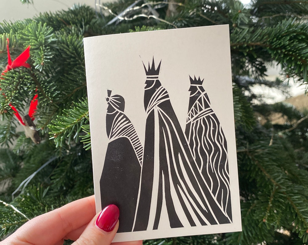 A greetings card is held in front of a christmas tree. The design features a three kings lino print